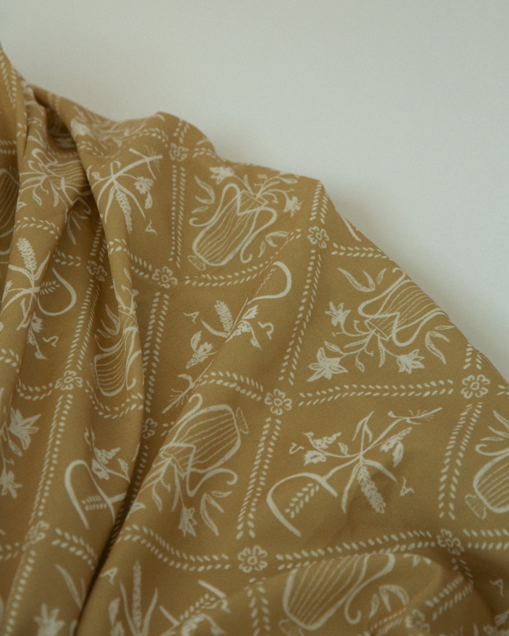A close-up of draped fabric in a warm tan or camel color with a repeating Catholic pattern, set against a plain off-white background.