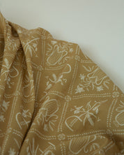 A close-up of draped fabric in a warm tan or camel color with a repeating Catholic pattern, set against a plain off-white background.