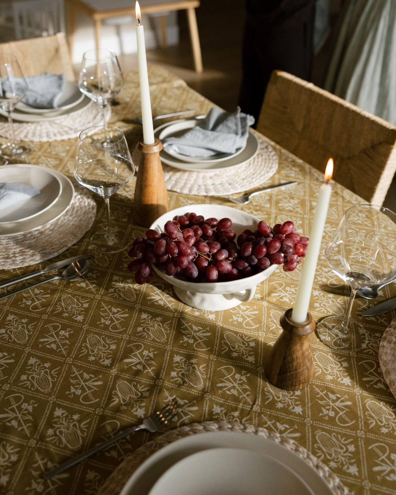 A styled dining table setting with a patterned olive green and beige tablecloth, white plates, gray napkins, lit taper candles in wooden holders, and a bowl of red grapes in the center. A person is partially visible in the background near wooden chairs.
