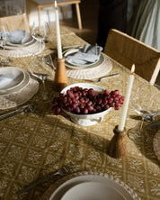 A styled dining table setting with a patterned olive green and beige tablecloth, white plates, gray napkins, lit taper candles in wooden holders, and a bowl of red grapes in the center. A person is partially visible in the background near wooden chairs.
