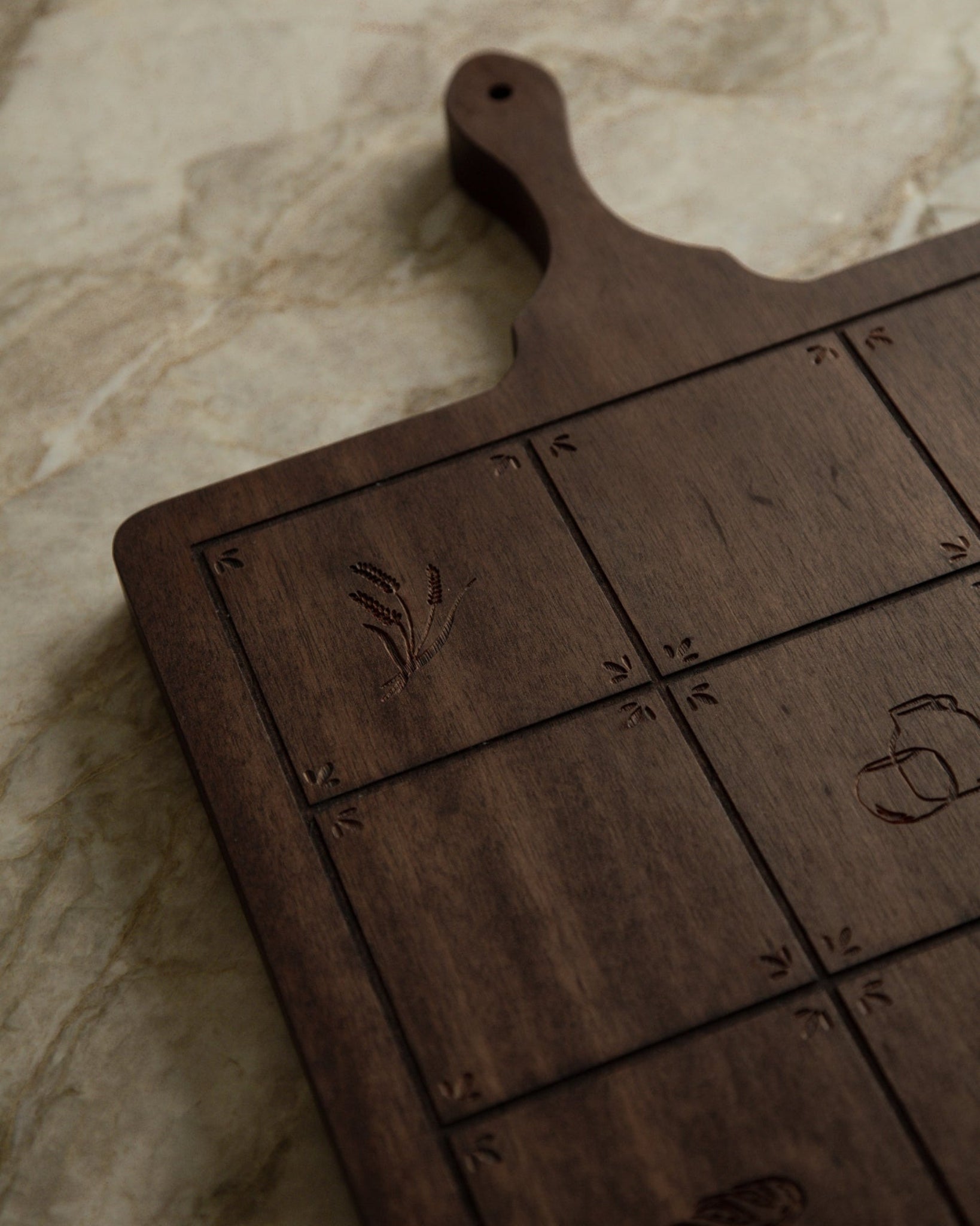 Dark brown wooden serving board with engraved grid sections and small Catholic icons, photographed on a light beige stone surface.