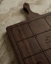 Dark brown wooden serving board with engraved grid sections and small Catholic icons, photographed on a light beige stone surface.