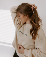 Woman wearing a beige sweatshirt with text, standing against a neutral background