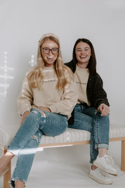 Two woman wearing beige sweater on bench with a light background