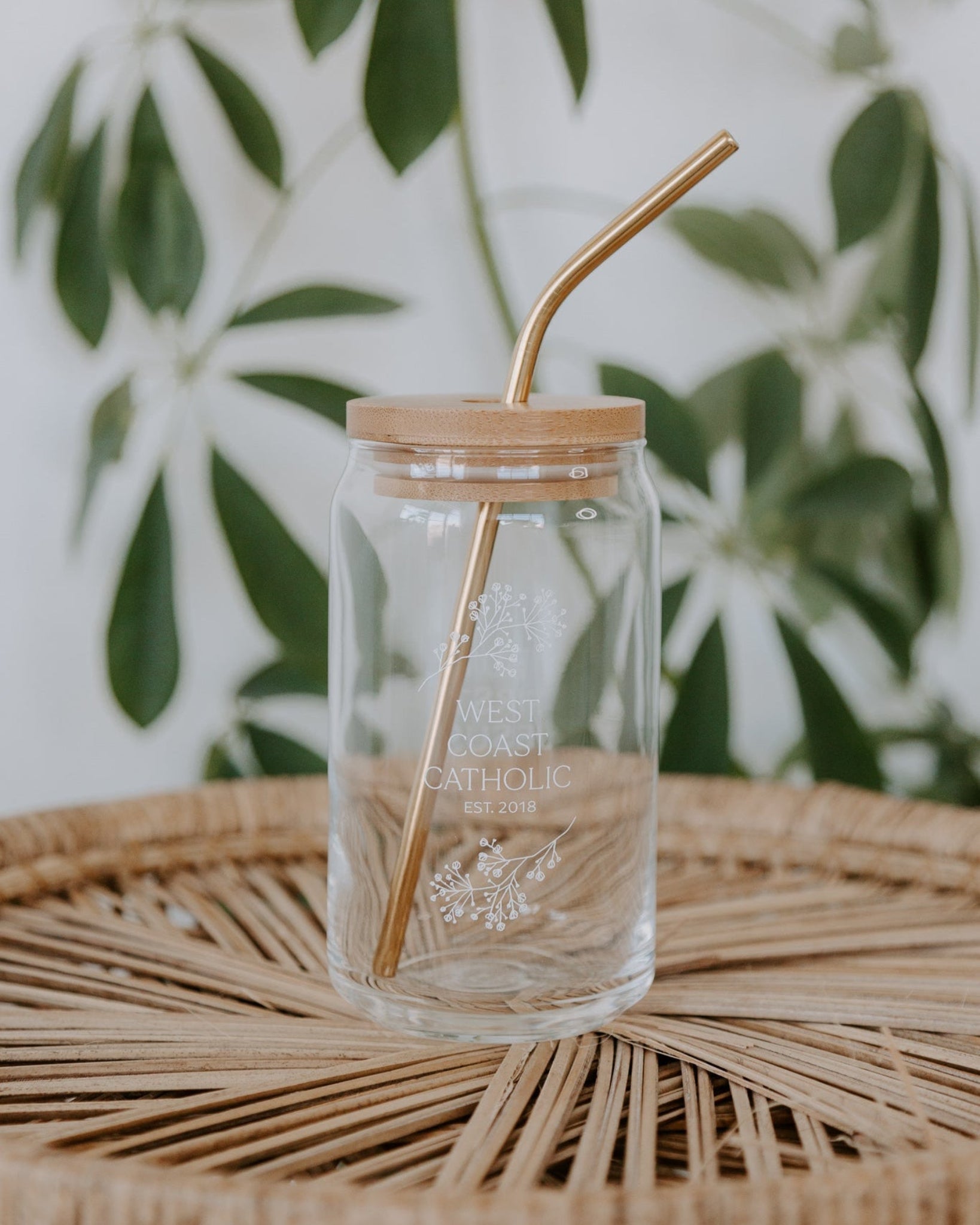 Clear glass jar with wooden lid and straw on a woven surface with green leaves in the background