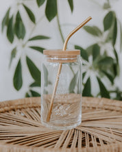 Clear glass jar with wooden lid and straw on a woven surface with green leaves in the background